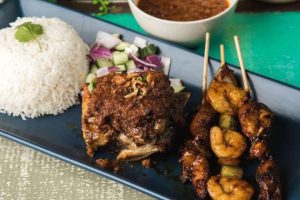 A mixed grill platter of beef ribs, chicken skewers and prawn skewers served with peanut sauce, cucumbers, red onions, and rice.